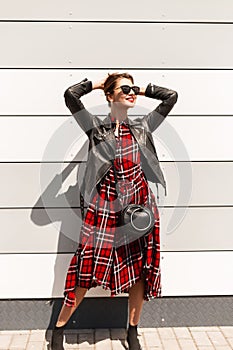 Happy young woman hipster with red lips in fashion black-red clothes in sunglasses straightens hairstyle standing on street near