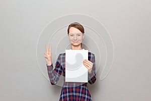 Happy young woman is holding blank paper sheet, showing victory sign
