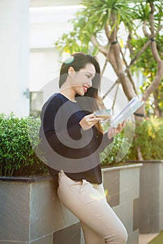 Happy young woman holding book fond of literature analyzing novel during leisure time on terrace of campus cafe in sunny day