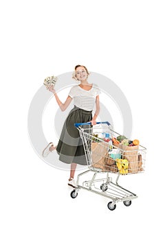 happy young woman holding dollar banknotes and standing with shopping trolley full of grocery