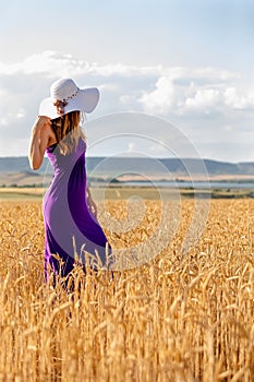 Happy young woman holding hat, walking in the golden wheat field. Sunset light. Summer nature.