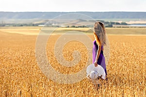 Happy young woman holding hat, walking in the golden wheat field. Sunset light. Summer nature.