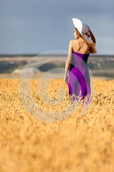 Happy young woman holding hat, walking in the golden wheat field. Sunset light. Summer nature.