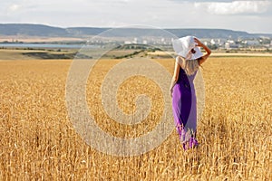 Happy young woman holding hat, walking in the golden wheat field. Sunset light. Summer nature.