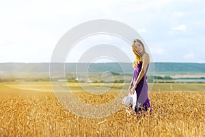 Happy young woman holding hat, walking in the golden wheat field. Sunset light. Summer nature.