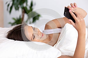Happy young woman holding phone lying in bed in morning