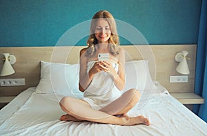 Happy young woman holding smartphone after bath with wrapped towel sitting on the bed in the morning