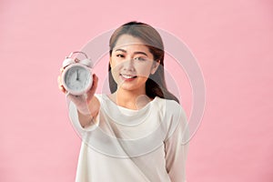 Happy young woman holding white alarm clock and waving hand isolated on pink