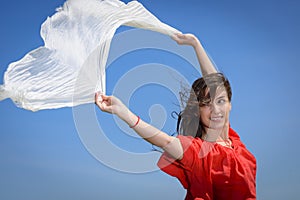 Happy young woman holding white scarf with opened arms expressing freedom, outdoor shot against blue sky