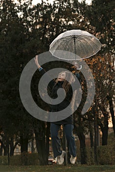 Happy young woman is jumping with transparent umbrella in hand. Girl with long curly hair in autumn park