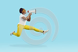 Happy young woman jumps in air and shouts through megaphone isolated on blue background