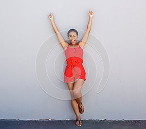 Happy young woman leaning against gray wall with arms raised