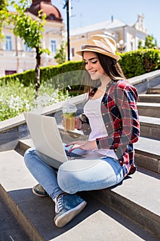 Happy young woman with limonade sitting on the city stairs and using laptop