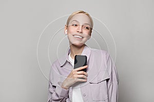 Happy young woman listening to music or podcast with headphones and holding smartphone on gray background