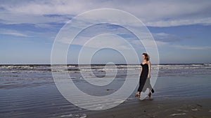 Happy young woman in long black dress walking on the beach with  golden retriever dog outdoors