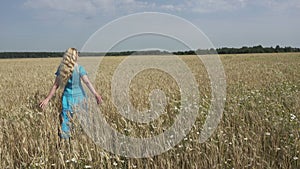 The happy young woman with a long fair hair and in a blue dress goes across the field of ripe wheat to sunny summer day