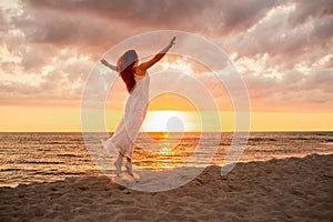 Happy young woman in a long white dress looking at the sunset on empty sand beach with her hands up. Freedoom, vacation,