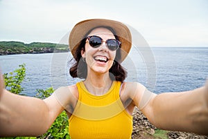 Happy young woman looking at camera taking selfie near the ocean during summer vacation.
