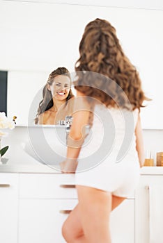 Happy young woman looking in mirror in bathroom