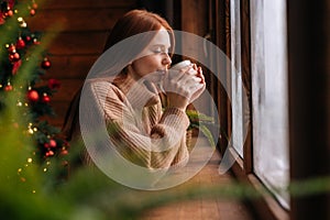 Happy young woman looking out of window and drinking hot coffee at home at Christmas time.