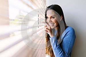 Happy young woman looking through venetian blinds on window