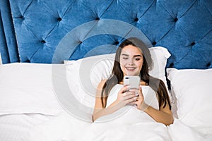 Happy young woman lying in bed and texting on smartphone at home bedroom