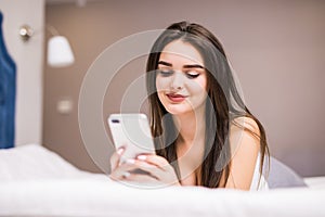 Happy young woman lying in bed and texting on smartphone at home bedroom