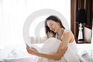Happy young woman lying in bed and texting on smartphone at home bedroom