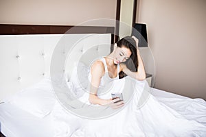 Happy young woman lying in bed and texting on smartphone at home bedroom