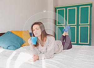 Happy young woman lying down on the bed with cup of tea and relaxing at home
