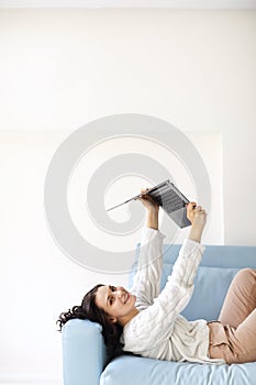 Happy young woman lying down on cozy sofa at home holding open laptop above her with hands up
