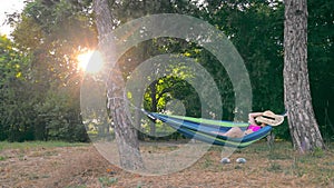 Happy young woman lying in a hammock in the park at sunset and bed hat on her face. Wonderful rest in the park on a