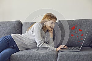 Happy young woman lying on sofa with laptop, video calling her boyfriend or chatting online