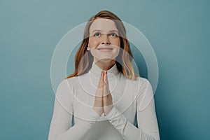 Happy young woman making wish while folded hands in prayer gesture, isolated on blue background