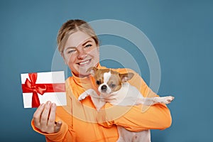Happy young woman in orange t-shirt with Chihuahua dog holds gift certificate  on blue background. People lifestyle