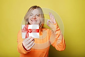 Happy young woman in orange t-shirt holds gift certificate isolated on yellow background. People lifestyle holidays concept