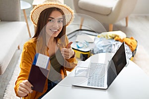 Happy young woman with passport and tickets using laptop, showing thumb up, recommending internet travel agency at home