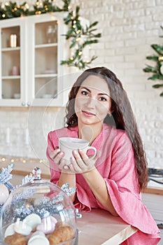 Happy young woman in pink dress with cup in Christmas decorated kitchen. beautiful brunette with cup of cocoa in kitchen with