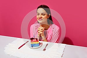 Happy young woman in pink shirt sitting at table with plate and birthday cake made of dishwashing sponges and candle and