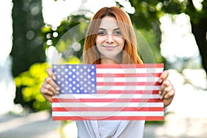 Happy young woman posing with USA national flag holding it in her outstretched hands standing outdoors in summer park. Pretty girl