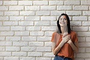 Happy young woman posing by wall putting hands over heart