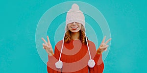 Happy young woman pulled hat over her eyes wearing white hat with pom pom, red knitted sweater on a blue background