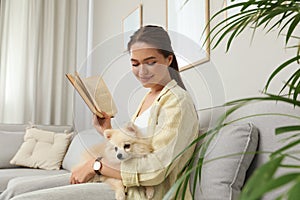Happy young woman reading book with cute dog on sofa in living room