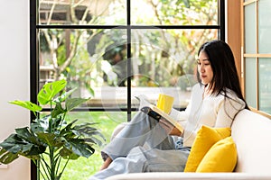 Happy young woman reading book on sofa at home.  Lifestyle freelance relax and chill drinking coffee in living room