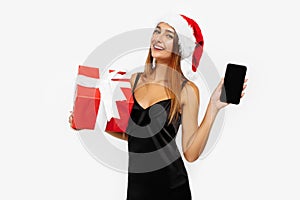 Happy young woman in red christmas hat holding gift box and showing blank mobile phone screen for copy space on white background