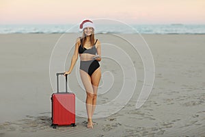 Happy young woman in red Christmas Santa Claus hat and bikini with suitcase on sand seashore shows hand for advertising