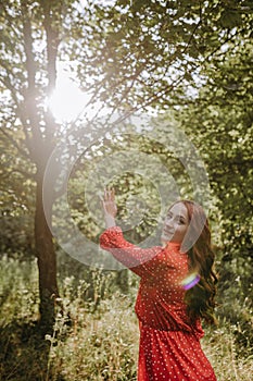 Happy young woman in red dress and with red long curly hair reaching out her hands to the sun in the forest