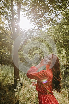 Happy young woman in red dress and with red long curly hair reaching out her hands to the sun in the forest