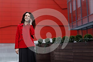 Happy young woman, in red jacket and skirt, talking on mobile phone and gesture with hand, over building background.