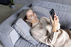 Happy young woman relax on comfortable couch at home texting messaging on smartphone, smiling girl use cellphone, browse wireless